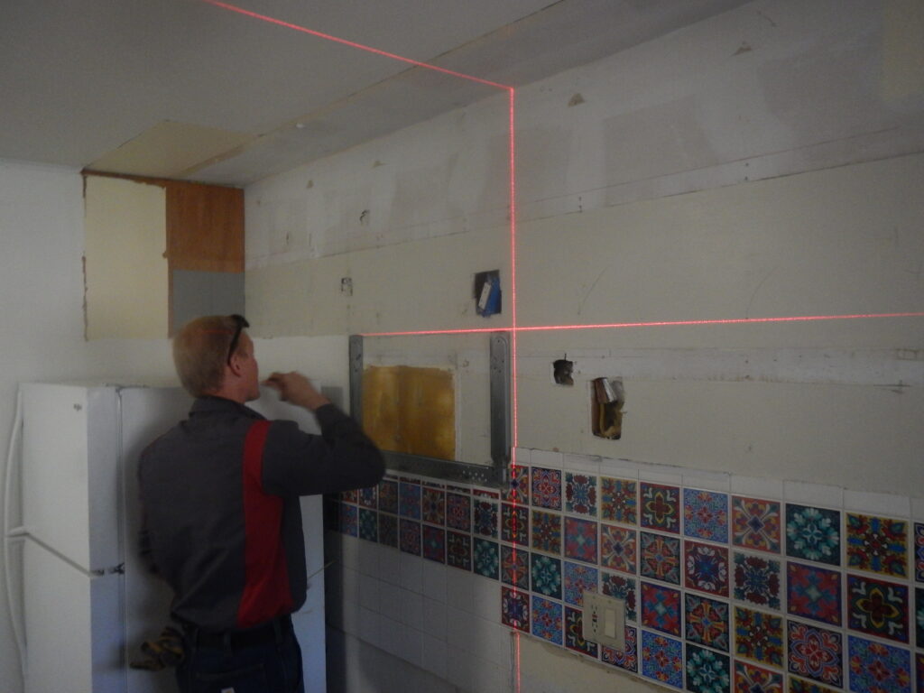 A red laser level projects straight lines to aid a handyman in hanging new upper kitchen cabinets.